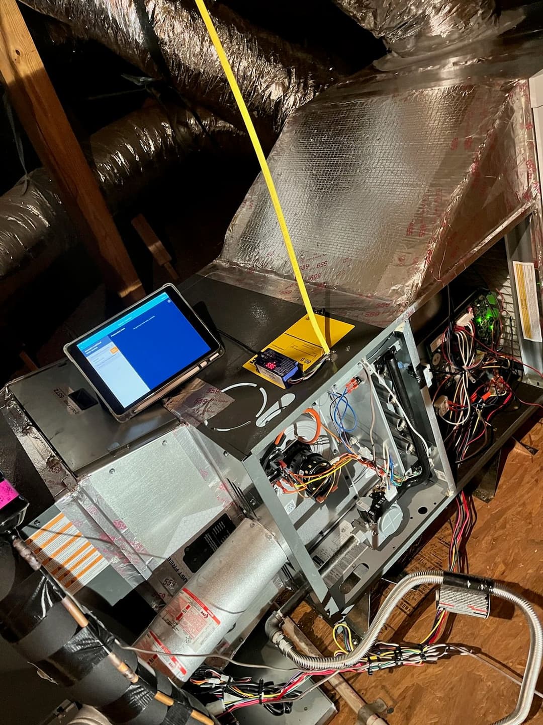 Tablet displaying HVAC system diagnostics in an attic with visible wiring and insulation.