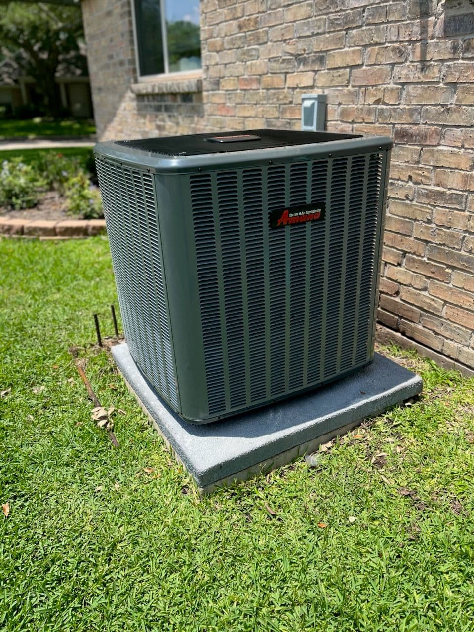 Air conditioning unit installed outside a brick house, sitting on a concrete slab in a grassy area.
