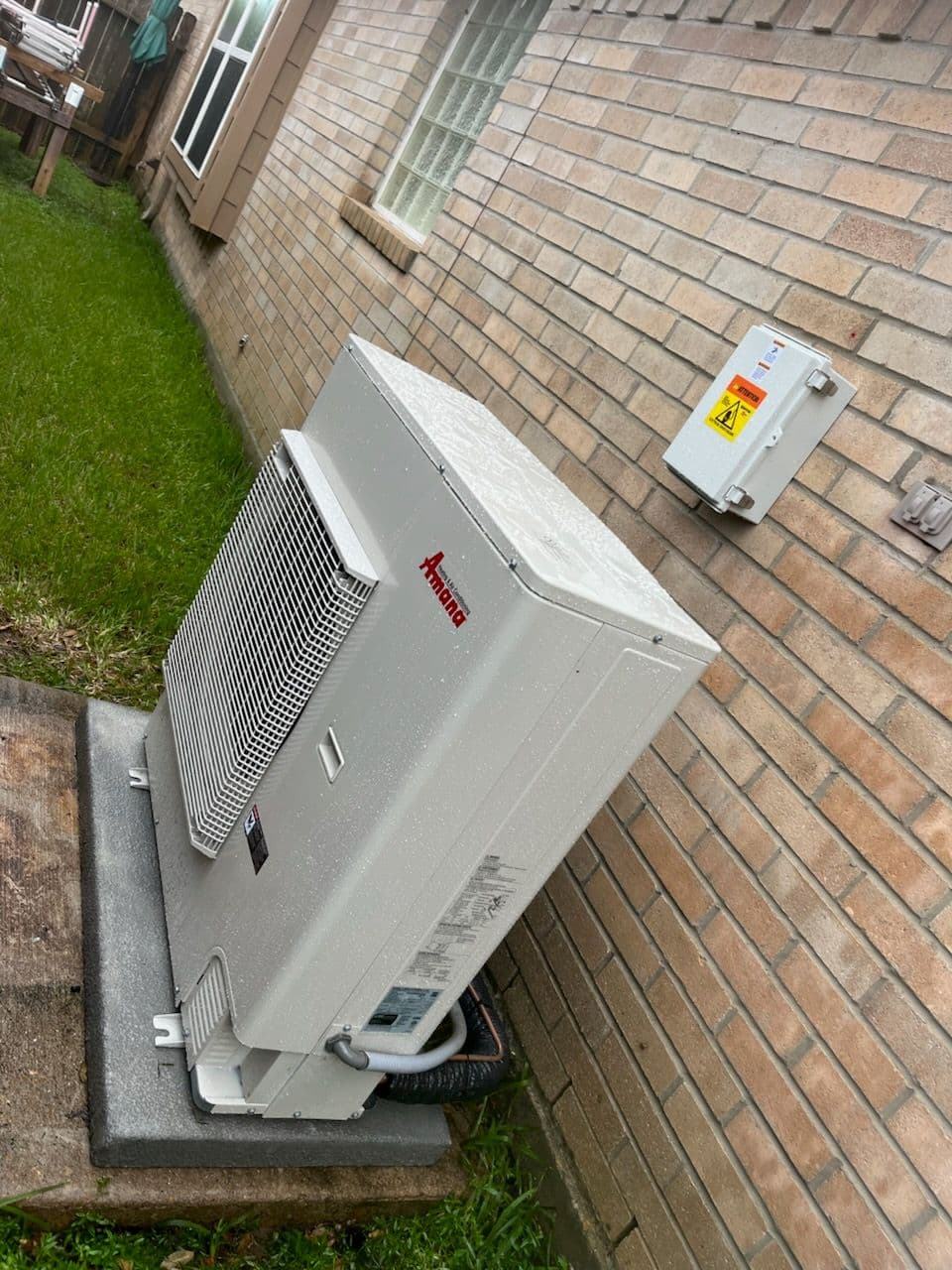 Air conditioning unit installed outside a home, on a grassy area next to a brick wall.