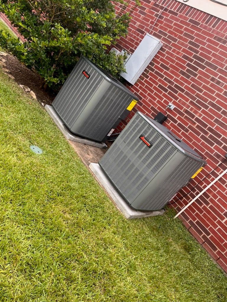 Two air conditioning units next to a brick wall with green grass nearby.