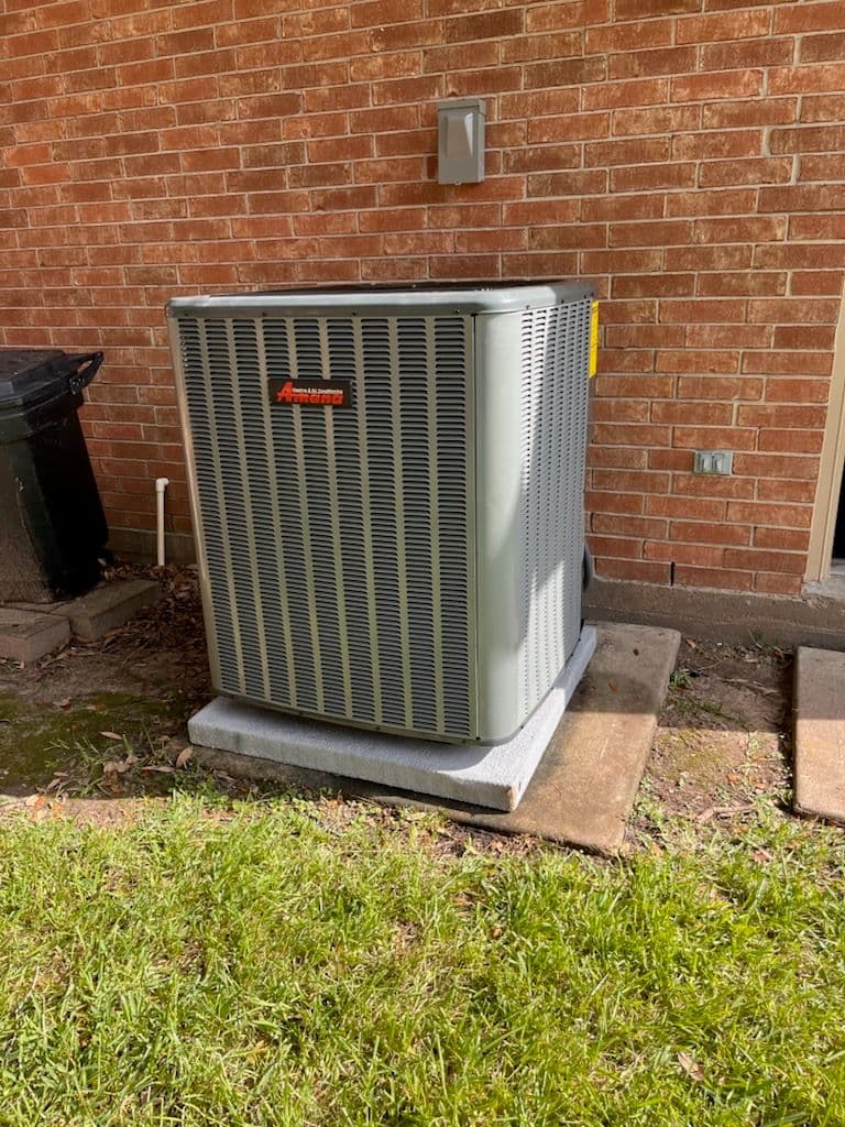 Air conditioning unit on a concrete base next to a brick wall and trash can.
