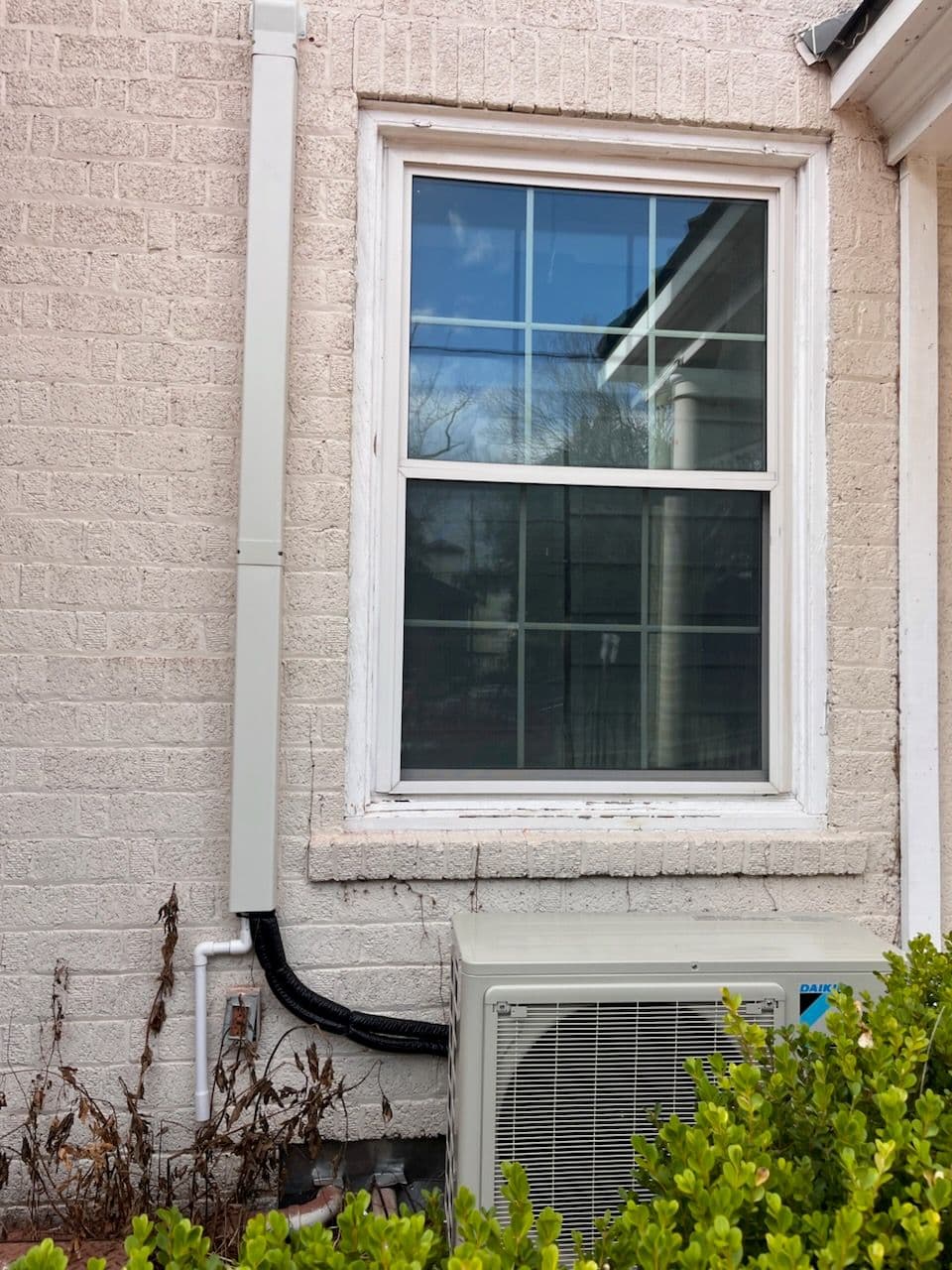 Window beside air conditioning unit on a textured brick wall with greenery below.