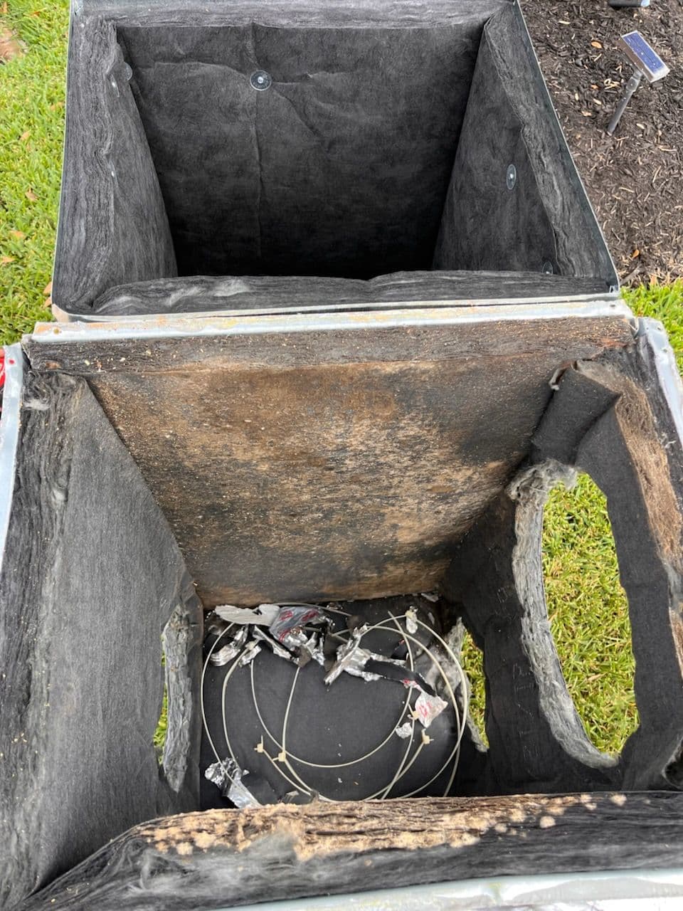 Empty, damaged recycling bins showing wires and debris inside on green grass.